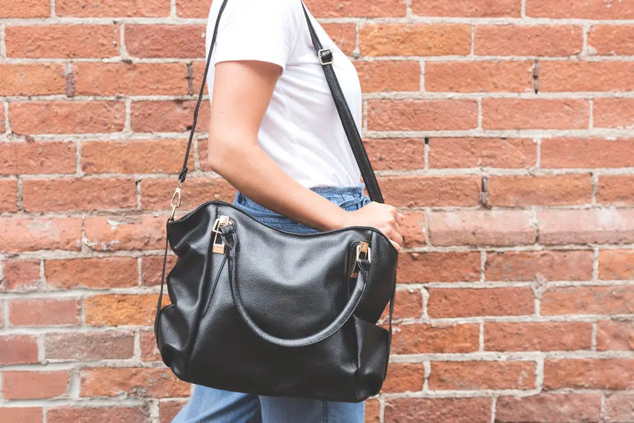 Black leather handbag with dual handles and shoulder strap, worn by person in white shirt and jeans against brick wall. Spacious design with gold-toned hardware, suitable for various carrying styles.